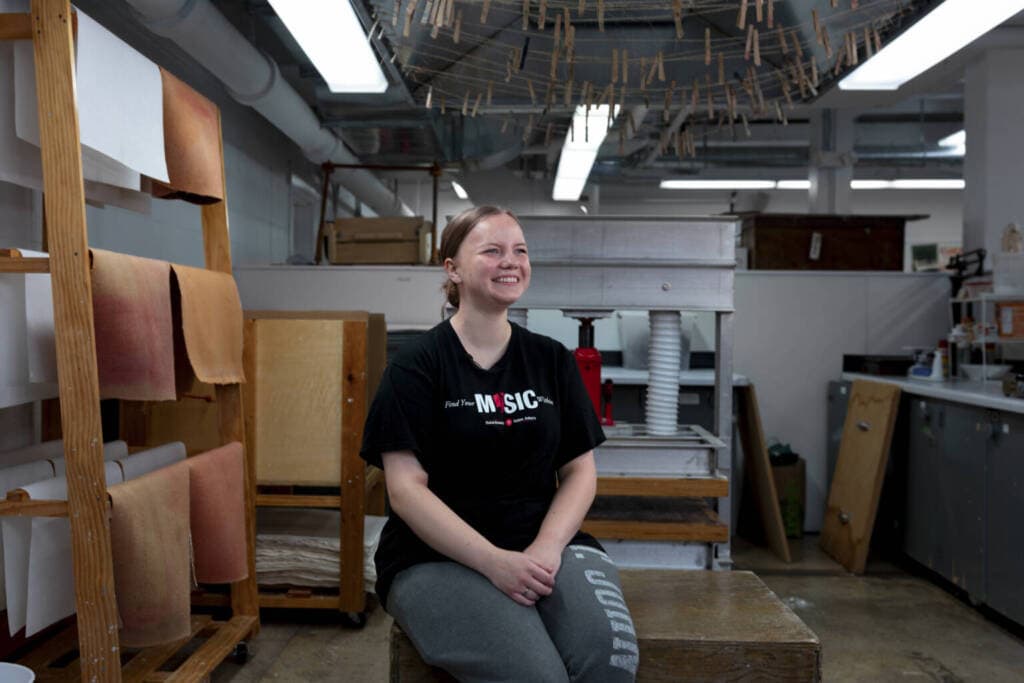 A smiling woman sits on a wooden bench in an art studio, wearing a black “MUSIC” T-shirt and gray pants. Shelves with rolled paper and clothespins hanging above are visible in the background.