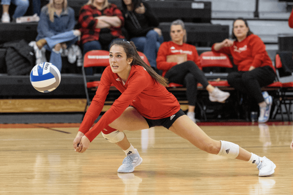 A volleyball player in a red jersey lunges forward with arms outstretched to bump the ball, while spectators watch from the bleachers in the background.