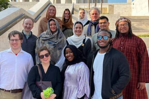 A diverse group of thirteen people pose together on outdoor steps in front of a modern white building with arches, under a blue sky with scattered clouds. Some wear sunglasses and traditional or casual clothing.