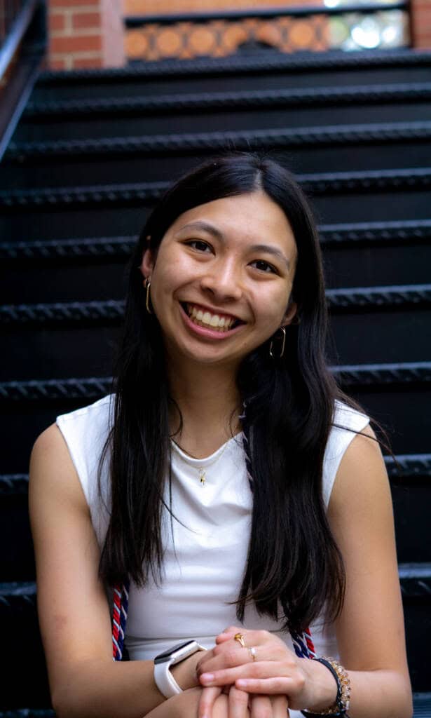 A young woman with long dark hair, wearing a white sleeveless top and gold jewelry, sits on black metal stairs, smiling at the camera with her hands clasped in front of her.