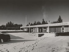 Black and white photograph of a modern, single-story building surrounded by snow. Footprints are visible leading to the entrance. Trees are visible in the background, and smoke is rising from a chimney on the right side of the building.