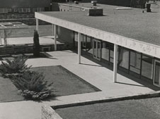 A black-and-white image of a modern building with a flat roof and large windows. The building has an overhang supported by white pillars and is surrounded by paved walkways and landscaped areas with small shrubs and grass patches.