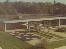 An aerial view of a single-story building with a flat roof and expansive windows on one side. In front of the building, there is a landscaped area with a geometric pattern of pathways and a modern pergola structure. Trees and another building are visible in the background.