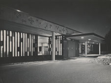 Black and white image of a modern building at night. The facade features vertical rectangular windows with varying lengths, allowing light to shine through. A covered walkway extends from the entrance, and decorative patterns adorn the upper part of the building.