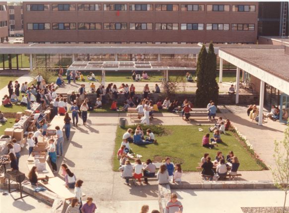 A lively outdoor scene on a university campus shows numerous students gathered in groups, sitting on benches, grass, and low walls. They are enjoying sunny weather near academic buildings with a mix of socializing and studying. Flowers and trees are visible in the background.