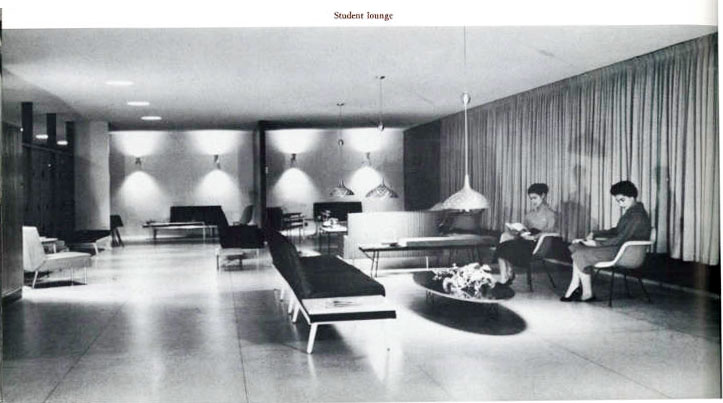 Black and white image of a "Student Lounge" featuring modern furniture, including long benches, a coffee table, pendant lights, and chairs. Two people are seated on the right side, reading. The space is spacious with curtains covering the back wall.