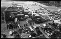 A black-and-white aerial view of a town showcasing a mix of residential buildings, institutional structures, and open fields. Straight roads with scattered vehicles intersect the town, and there are areas of dense greenery and a few larger buildings, possibly schools or factories.