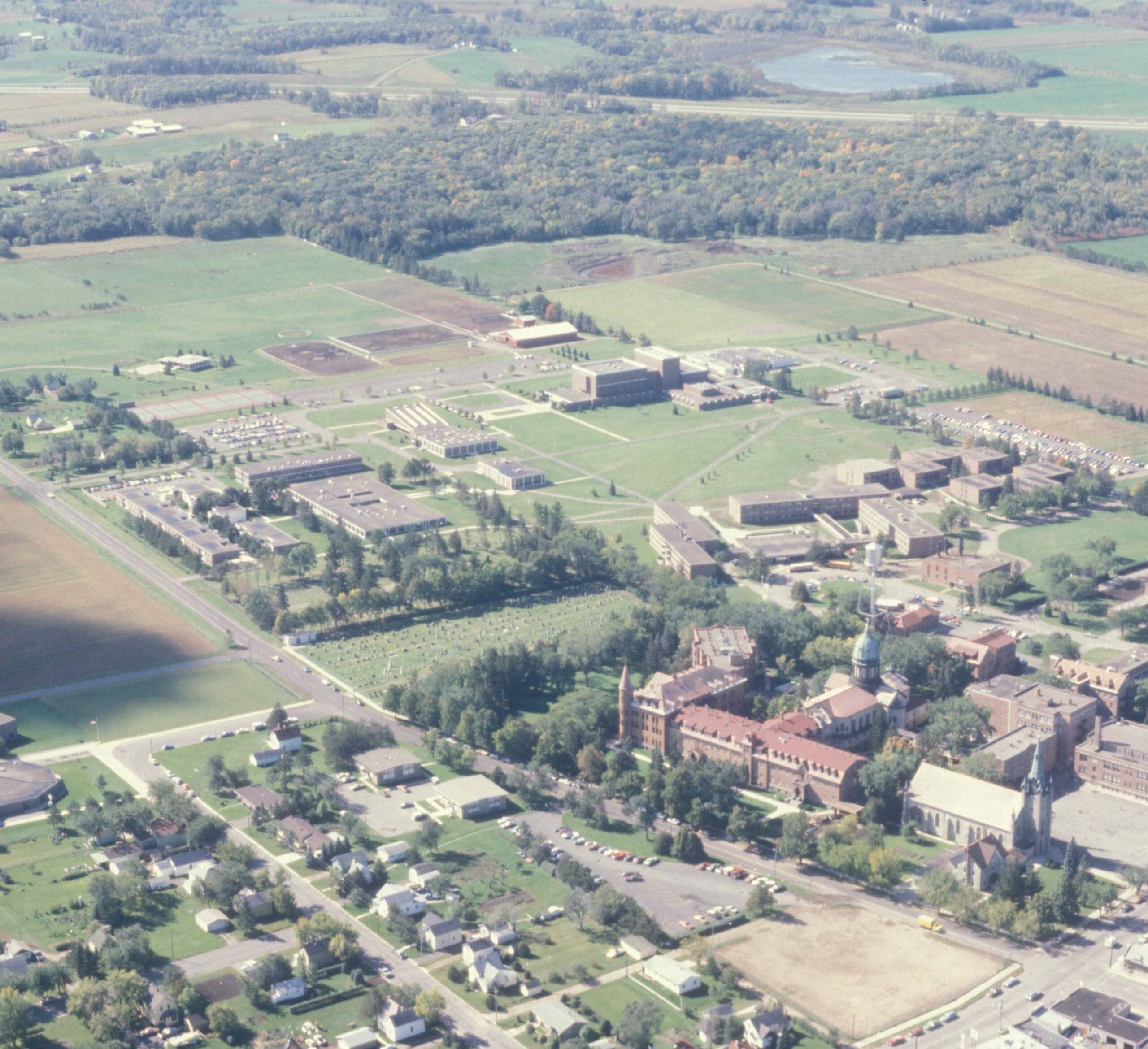 Aerial view of a large campus with multiple buildings scattered across green fields. The image also shows a mix of residential areas, roads, and surrounding countryside with trees and fields in the distance.