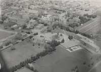 A black-and-white aerial photograph of a university campus with multiple buildings surrounded by trees and green lawns. The campus includes a sports field, various academic buildings, and a central area with tree-lined paths. The cityscape is visible in the background.