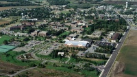 Aerial view of a suburban area featuring residential houses, commercial buildings, and verdant greenery. Roads and pathways connect various structures, with fields and trees scattered throughout the landscape.