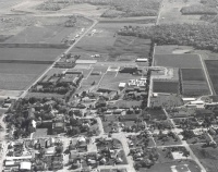 Aerial black and white photograph of a sprawling rural landscape featuring scattered buildings, farmland, and a network of roads. The scene includes residential areas, agricultural fields, and clusters of structures indicating possible institutional or industrial facilities.