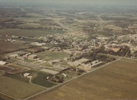 Aerial view of a small town surrounded by fields and farmland. The town has a mix of residential and commercial buildings clustered together. Roads intersect the area, and the landscape stretches out towards a hazy horizon under a clear sky.