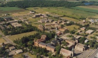 Aerial view of a sprawling academic campus with multiple brick buildings surrounded by green fields and scattered trees. The campus is situated near a forested area and several open fields. Pathways and roads interconnect the buildings. The sky is clear.