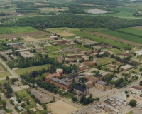 An aerial view of a small town with a mix of buildings, including homes, schools, and businesses, surrounded by green fields and trees. The layout shows a combination of urban and rural elements with roads connecting the various structures.