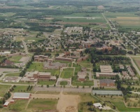 Aerial view of a large campus with numerous buildings surrounded by green spaces and trees. Roads connect various sections of the campus, with fields and distant urban areas in the background. The image shows an organized, expansive layout.