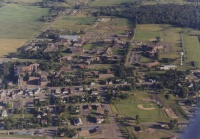 Aerial view of a small town surrounded by fields and greenery. There are various buildings, roads, and a baseball field in the center. The landscape is largely rural with scattered trees and agricultural areas.
