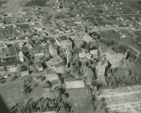 Aerial view of a town featuring a mix of residential, commercial, and industrial buildings. The landscape includes numerous houses, larger buildings likely to be businesses or institutions, and some open fields. Roads and vehicles are visible, and the setting appears serene.