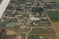 An aerial view of a small town surrounded by fields. The town features several buildings, roads, and green spaces. The landscape includes a mix of developed and undeveloped areas, with a noticeable large white-roofed building at the center.