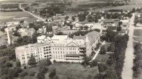 A black and white aerial view of a large building complex surrounded by trees and fields. The complex consists of multiple connected structures with various roof levels and architectural details. Roads and smaller buildings can be seen in the distance.