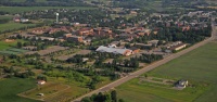 An aerial view of a small town surrounded by green fields and scattered trees. The town features a mix of residential and commercial buildings, with a notably large white-roofed structure in the center. Roads and pathways cut through the landscape.