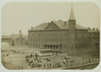 A sepia-toned photograph of a large, multi-story brick building with a tall, pointed tower. People are gathered outside, and a streetcar is parked in front. Additional smaller buildings are visible in the background. The image has a vintage appearance.