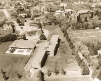 A sepia-toned aerial photograph of a campus featuring several buildings arranged around open green spaces. The foreground has a few large structures, while more buildings are scattered in the background with tree-lined pathways connecting them.