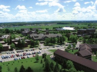 An aerial view of a sprawling campus with multiple buildings surrounded by greenery and parking lots filled with cars. The background features open fields under a blue sky with scattered clouds.