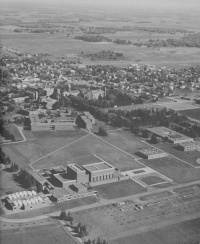 Aerial view of a mid-20th century town with several large buildings surrounded by open fields. The small town consists of clustered residential homes amidst a grid of streets, with some larger brick buildings and a parking lot in the foreground.