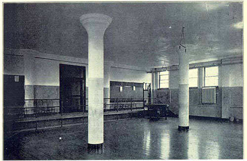 A black and white photo of a large, empty room with two prominent white pillars. The room has wooden floors and windows on the right side. A low railing is seen on the left side of the image. Lighting appears to be from natural sources.