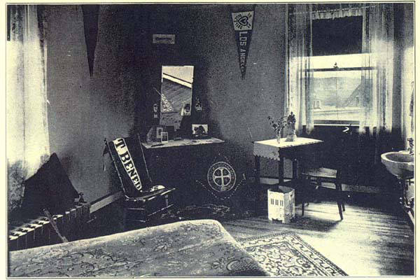 A vintage photograph of a simply furnished dorm room. The space includes a bed, a small table with two chairs, a desk with a mirror, a rocking chair with a blanket, and a small sink. Several pennants and framed pictures decorate the walls.