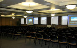 A large conference room with rows of empty black chairs facing two projection screens at the front. The room has a tray ceiling with recessed lights and round hanging fixtures. A podium stands near the screens, and there's a windowed wall in the background.