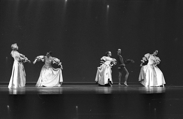 A group of dancers performing on stage in a dramatic pose. They are wearing long, flowing costumes, and the lighting creates a mysterious atmosphere. The background is dark, emphasizing the elegance and movement of the dancers.