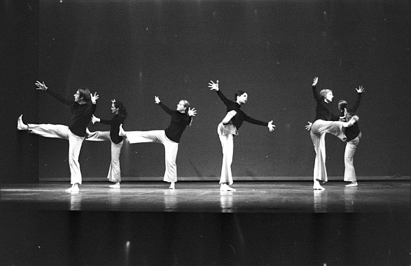 A group of six dancers perform on stage, each striking a graceful, synchronized pose. They wear dark tops and light pants, extending their arms and legs in unison against a plain backdrop. The scene is captured in black and white.