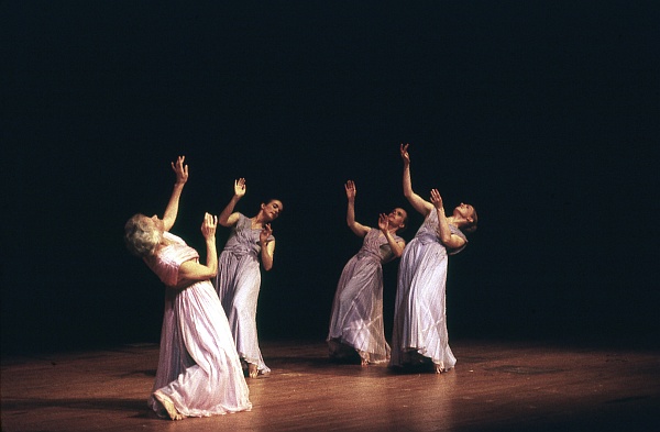 Four dancers in flowing dresses perform on a dimly lit stage, each in a graceful pose with arms extended upwards. The scene conveys an elegant and expressive performance.