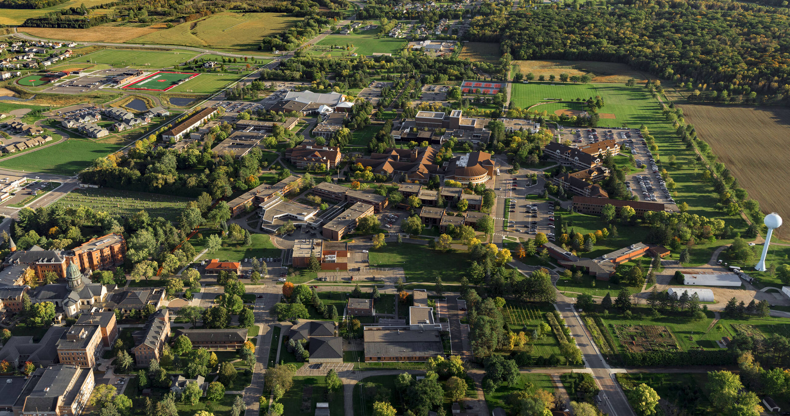 Aerial view of a rural area with a mix of buildings, trees, and open fields. A cluster of large buildings is centrally located, surrounded by roads and greenery. A water tower is visible in the lower right corner, and a sports field is in the upper left.