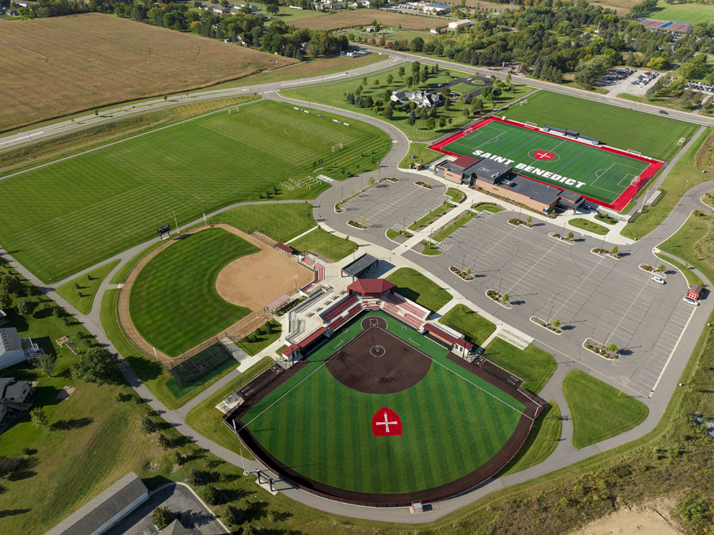 Aerial view of a sports complex with a football field labeled "SAINT BENEDICT," a baseball diamond, and a smaller field. There are parking lots and surrounding green areas, situated in a rural landscape with roads and trees in the background.