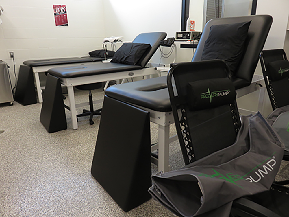 A room with several cushioned therapy tables and chairs, each equipped with pillows. A folded Recovery Pump device is visible beside the chairs. The floor is carpeted and the walls are white, giving the space a clinical feel.