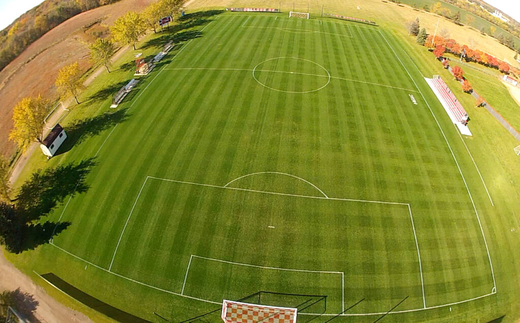 Aerial view of a well-maintained soccer field with marked white lines, goals at each end, and surrounding autumn foliage of trees in various shades of green, yellow, and orange. The field is bordered by a dirt path.