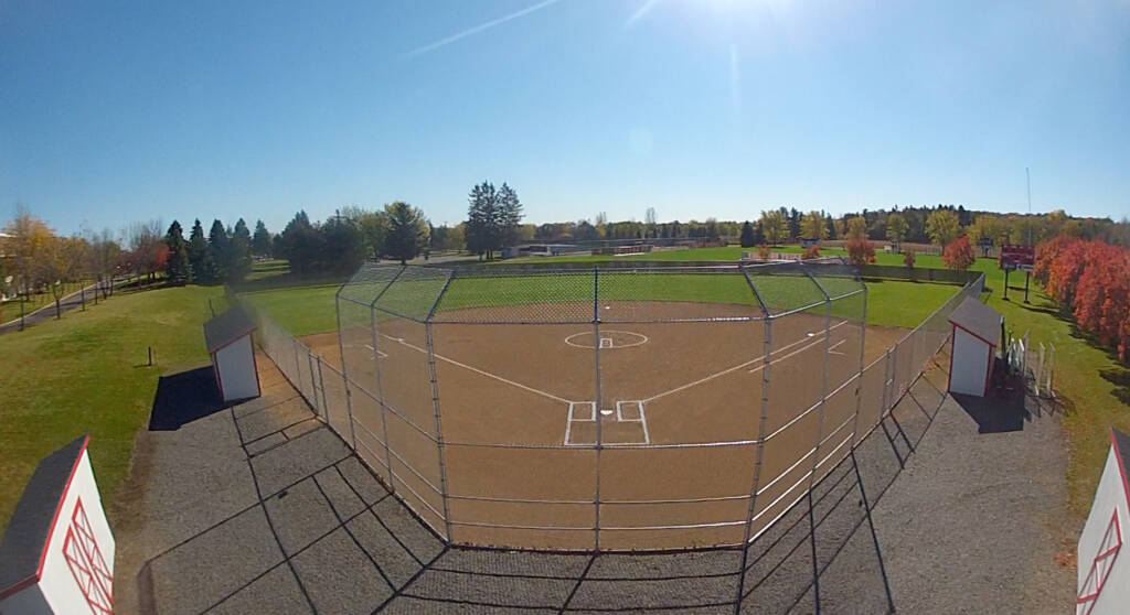 Aerial view of an empty baseball field on a sunny day. The diamond is centrally located, surrounded by green grass and trees in the background. The sky is clear blue, casting shadows on the field.