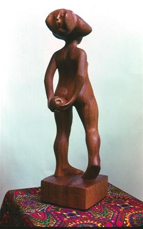 A wooden sculpture of a standing abstract human figure with an oversized, angled head and elongated limbs, holding an object near its waist. The statue is displayed on a colorful patterned cloth.