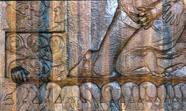 A carved relief depicts a large central figure's legs and hands, several smaller human figures to the left, and a row of animals along the bottom, all intricately detailed in a brown, textured surface.