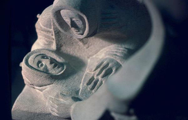 A close-up of a stone sculpture depicting two figures in robes, one lying down with hands folded and a peaceful expression, while the other leans over, gazing down with a gentle, caring demeanor.