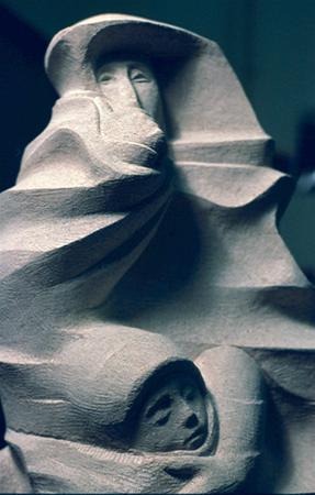 Abstract stone sculpture featuring two human figures; one stands draped in flowing, angular robes, partially obscuring their face, while a smaller, hooded figure appears nestled at the base, both rendered with minimal facial details.