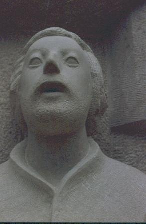 A close-up photo of a stone statue depicting a person with an open mouth and blank, rounded eyes, wearing a simple robe and set against a rough stone background.