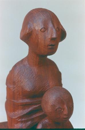 A wooden sculpture depicting two abstract human figures, one larger and one smaller, with simple facial features and rounded shapes, set against a plain background.