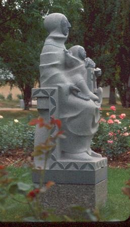 A stone statue of a seated figure holding a child on their lap, set outdoors in a garden with green grass, blooming flowers, and trees in the background.