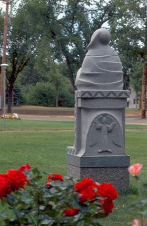 A gray stone statue stands in a grassy park, with a carved angel on its base. Red roses bloom in the foreground, and trees are visible in the background.