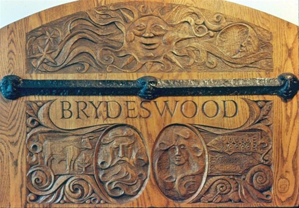 A wooden panel intricately carved with the word "BRYDESWOOD," a sun with a face, a horse, a ship, a woman's portrait, and a house, divided by a horizontal metal bar.