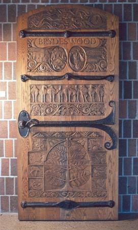 Ornate wooden door with intricate carvings, including people, animals, leaves, and the words "BRYDES WOOD" at the top, set against a brick wall. Black iron hinges and handle decorate the door.