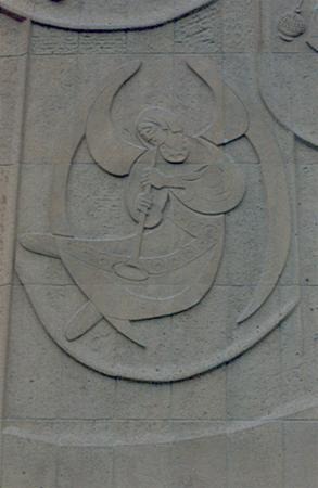 Stone relief carving depicting a figure in flowing robes holding a staff or rod, set within an abstract, oval-shaped design on a textured wall.